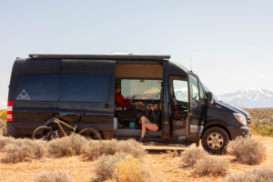 Woman taking photos from the door of a black sprinter van with snow covered peaks in the background.