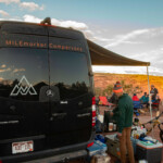 a man cooking on a camping stove under the awning of a campervan in the utah desert